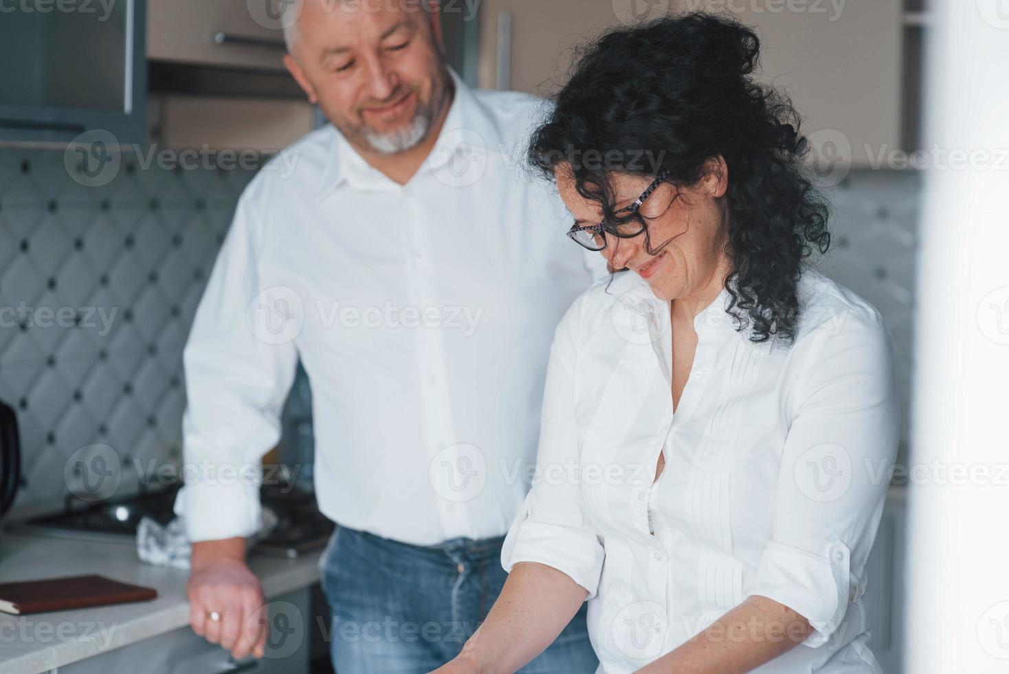 tendo bom humor. homem e sua esposa de camisa branca preparando comida na cozinha usando legumes foto