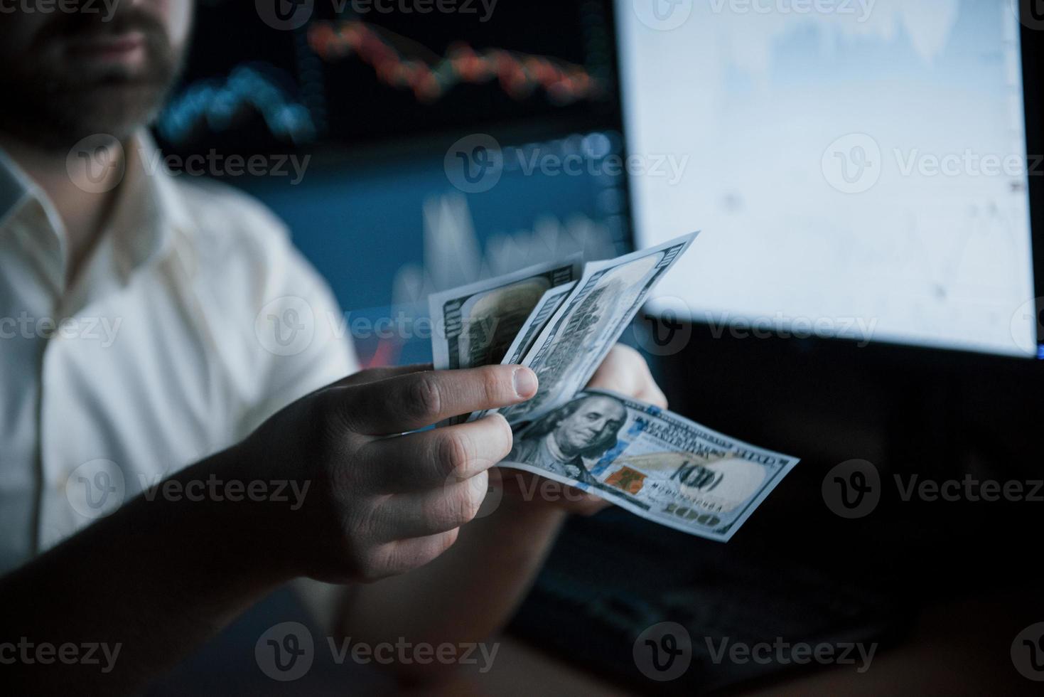 é dia de pagamento. homem barbudo de camisa branca conta dinheiro no escritório com várias telas de computador em gráficos de índice foto