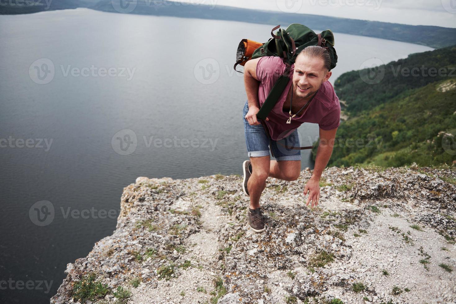 viajante com mochila sentado no topo da montanha, apreciando a vista acima da superfície da água. viajando ao longo das montanhas e da costa, conceito de liberdade e estilo de vida ativo foto