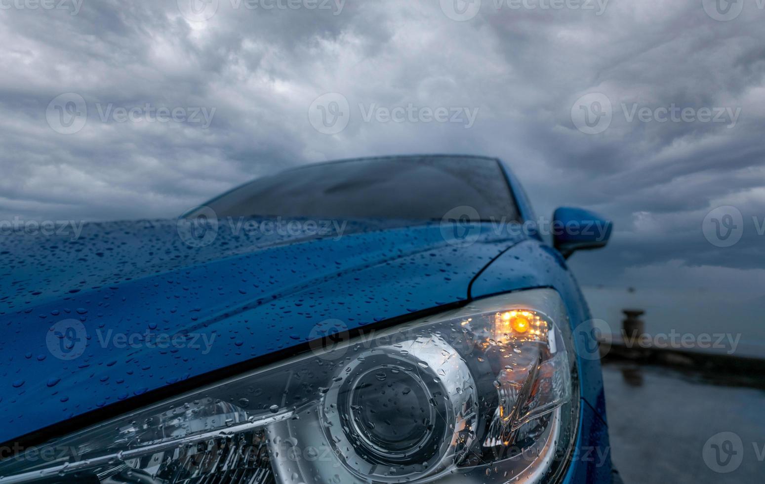 foco seletivo nos faróis do carro suv de luxo azul estacionado na estrada de concreto ao lado da praia do mar em dia chuvoso com céu escuro de tempestade. gotas de chuva no carro suv azul com farol aberto. viagem de viagem. foto