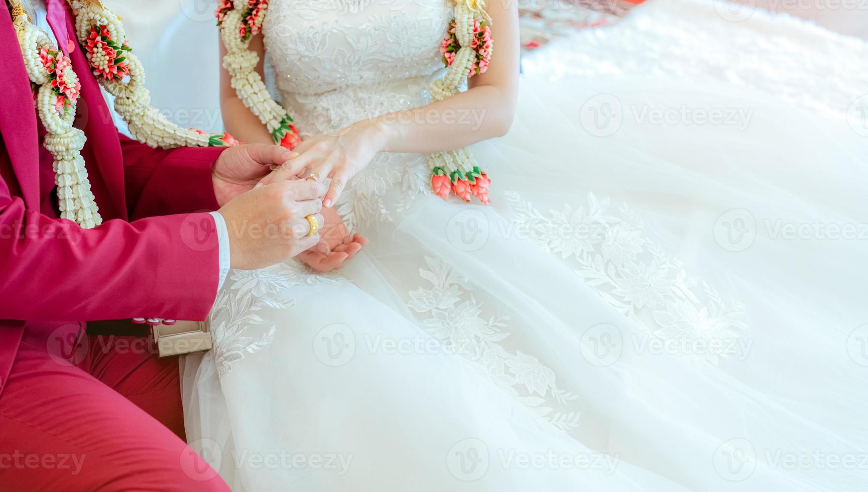 noivo colocou anel de diamante de casamento no dedo da noiva para proposta no dia da cerimônia de casamento. noiva e noivo em vestido de noiva branco e terno vermelho e guirlandas de casamento. casamento ou matrimônio. começar a vida de casal. foto