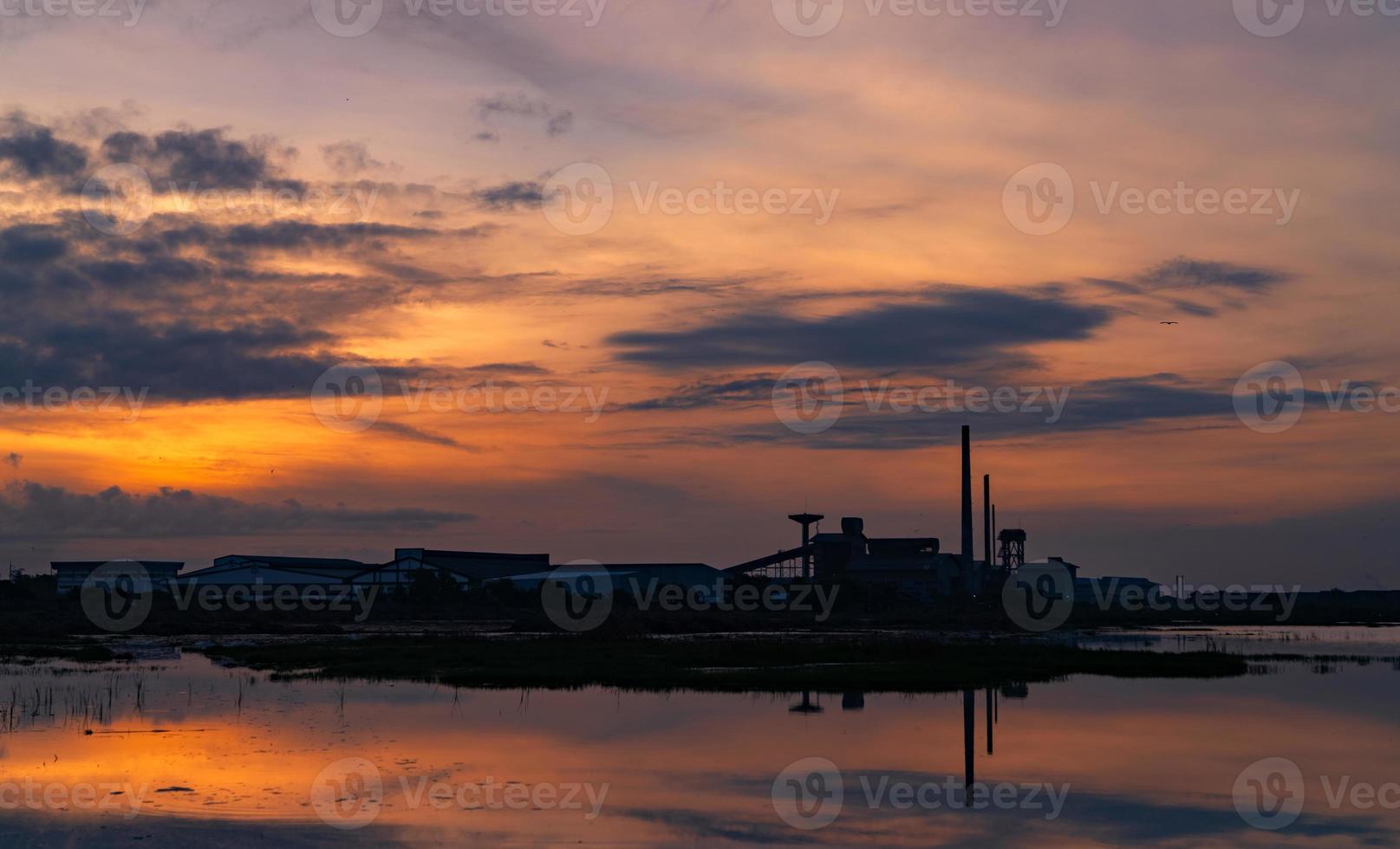 paisagem de edifícios da indústria fabril com reflexo azul escuro e laranja do céu do sol na água do rio. edifício do armazém à noite. ambiente limpo em torno da fábrica. fábrica fechada. foto