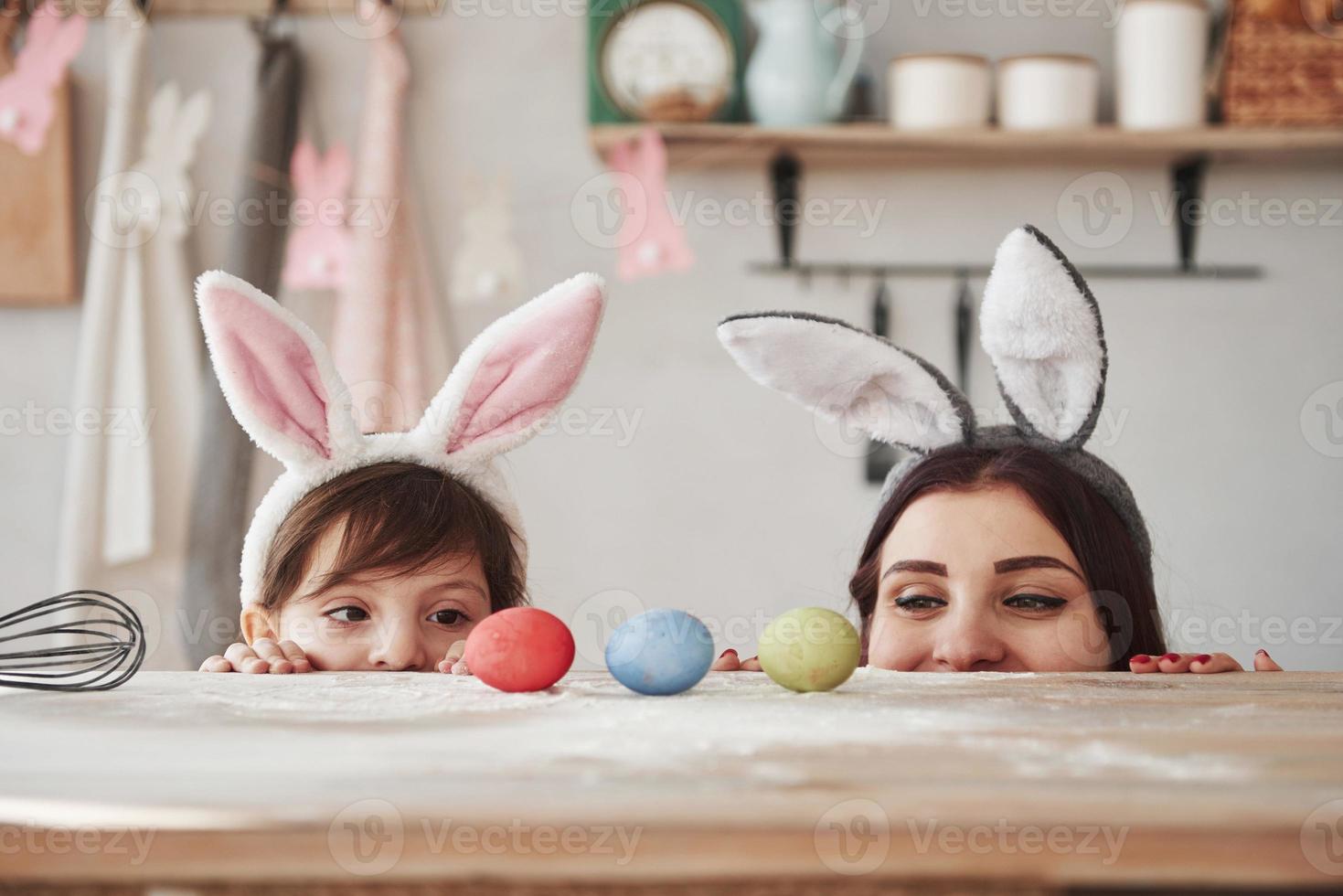 olha para os ovos. mãe e filha em orelhas de coelho na páscoa se divertem na cozinha durante o dia foto