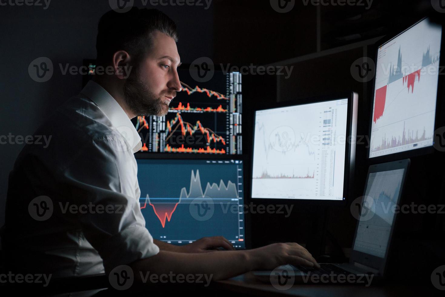 não pare seu trabalho mesmo à noite se quiser algum dinheiro extra. homem barbudo de camisa branca trabalha no escritório com várias telas de computador foto