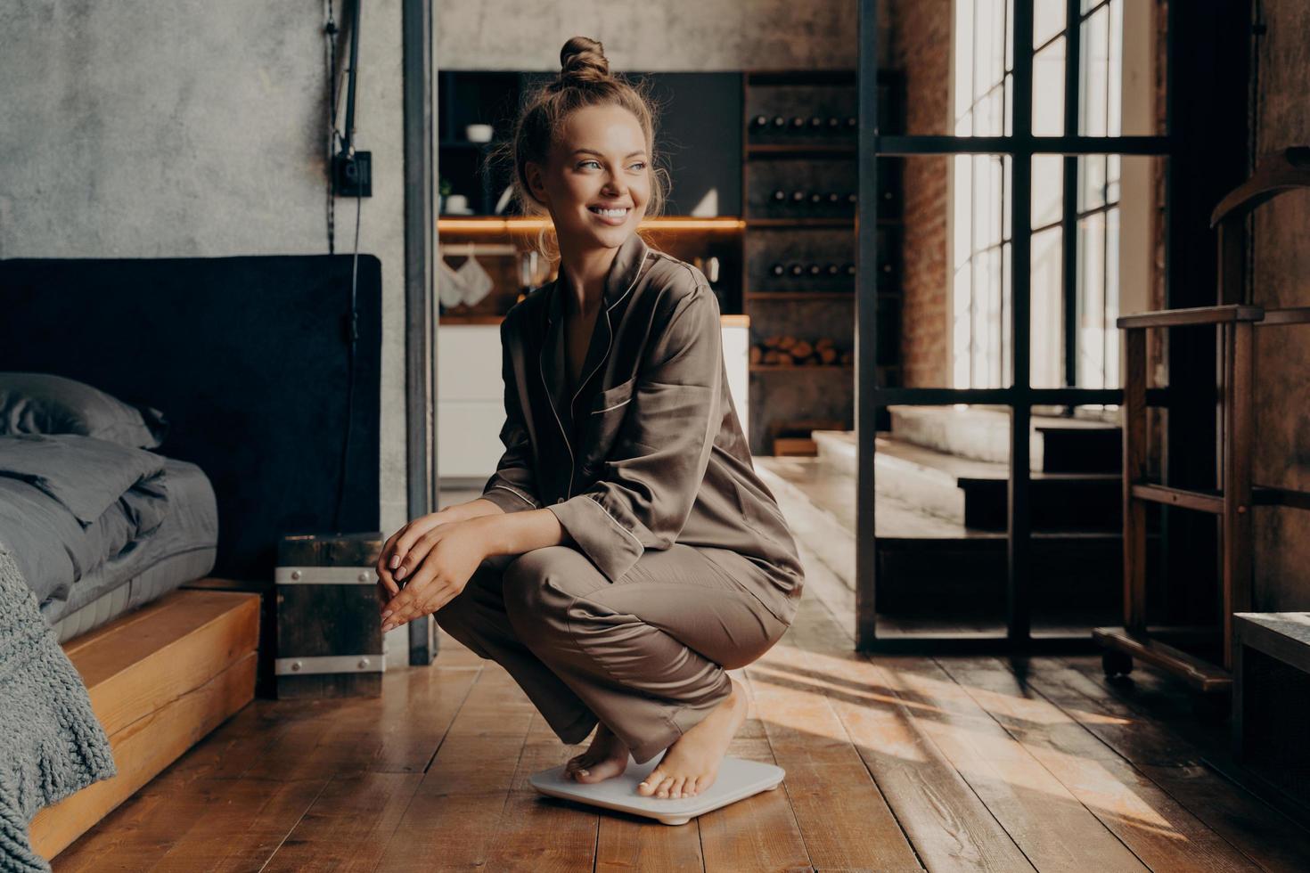 imagem do alegre modelo satisfeito jovem em pijama aconchegante de cetim agachado sobre balanças com um largo sorriso no quarto moderno foto