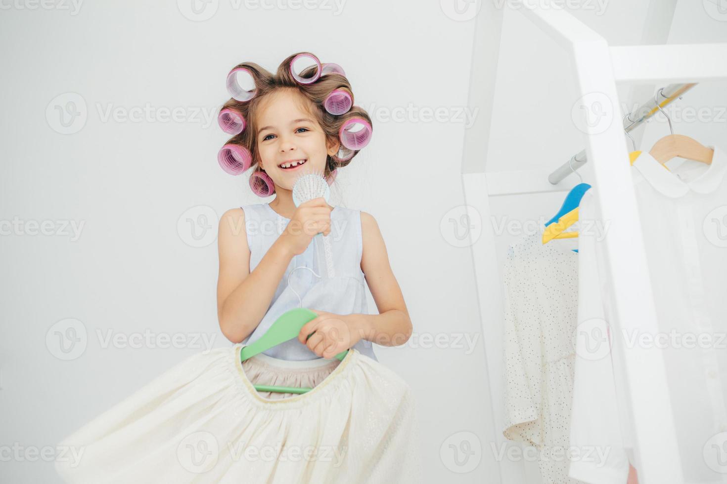 criança feliz do sexo feminino segura pente e vestido festivo em cabides, tem bobes no cabelo, se prepara para festa de aniversário com amigos, cuida de sua beleza e aparência, isolada no fundo branco foto