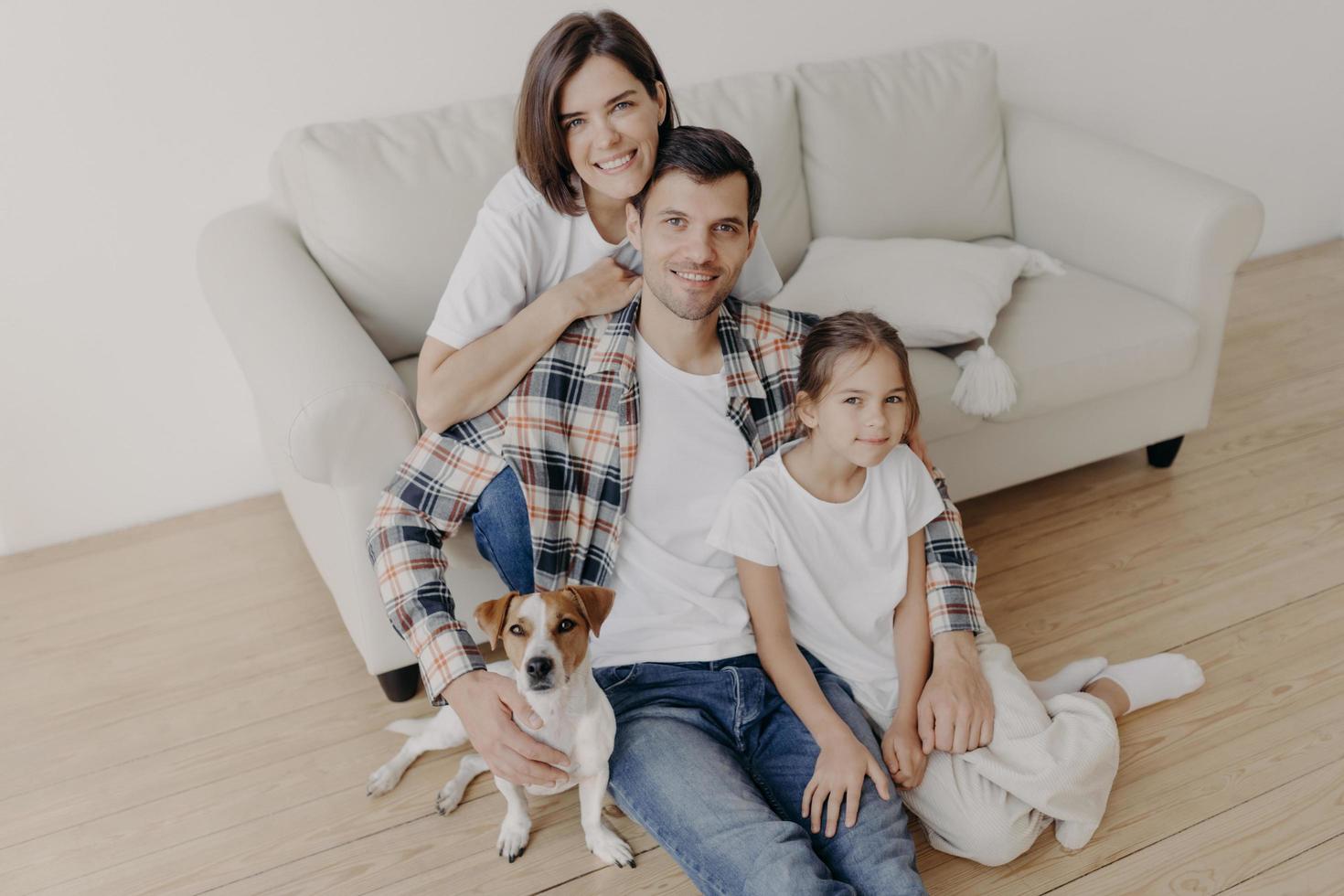 vista superior de membros da família felizes posam em uma sala espaçosa perto de um sofá confortável, poses de cachorro favorito perto, aproveite o tempo livre, sente-se no chão, sorria positivamente. pai, mãe e filho passam tempo em casa foto