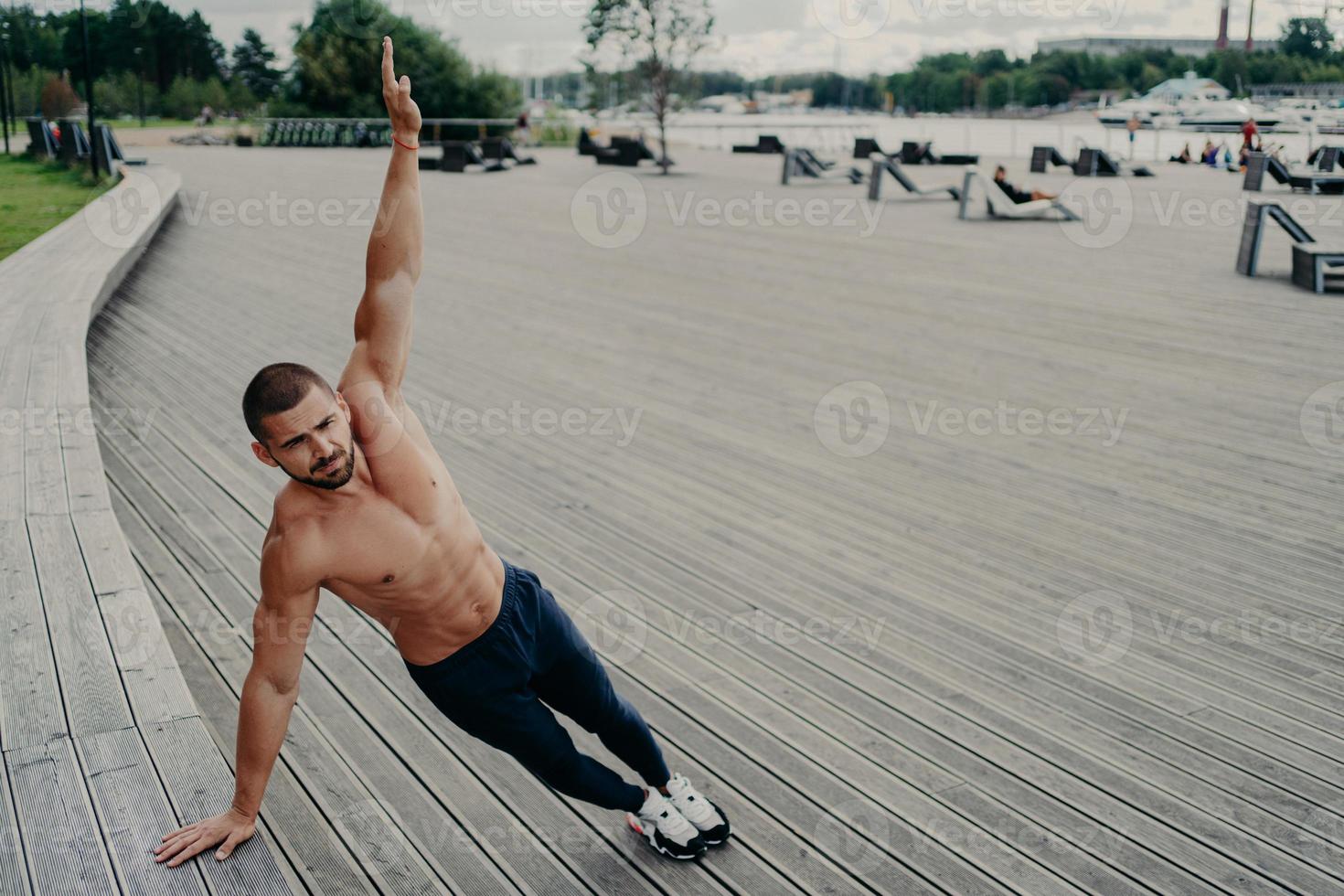 auto determinado homem musculoso vestido com roupas esportivas fica na prancha lateral, mantém o braço levantado, tem treino ao ar livre durante o dia de verão. esporte, estilo de vida saudável, conceito de força e resistência. foto