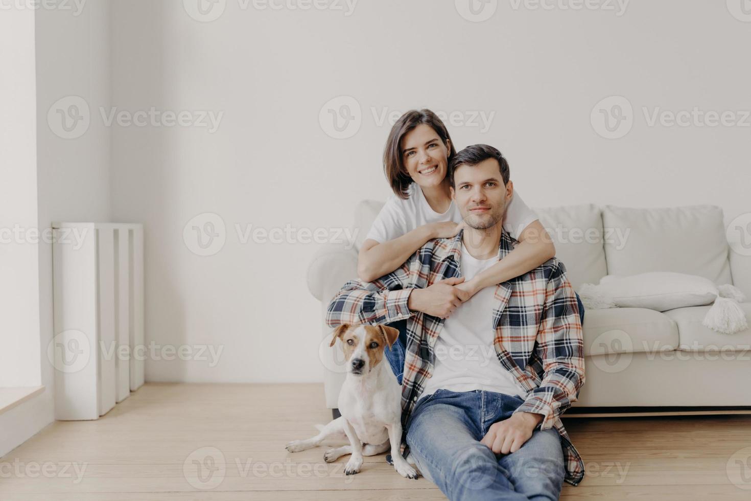 retrato de esposa afetuosa abraça marido que se senta no chão perto do sofá, seu animal de estimação favorito posa para a câmera, faz foto, estando em casa, compra apartamento novo, posa na sala de estar com paredes brancas foto