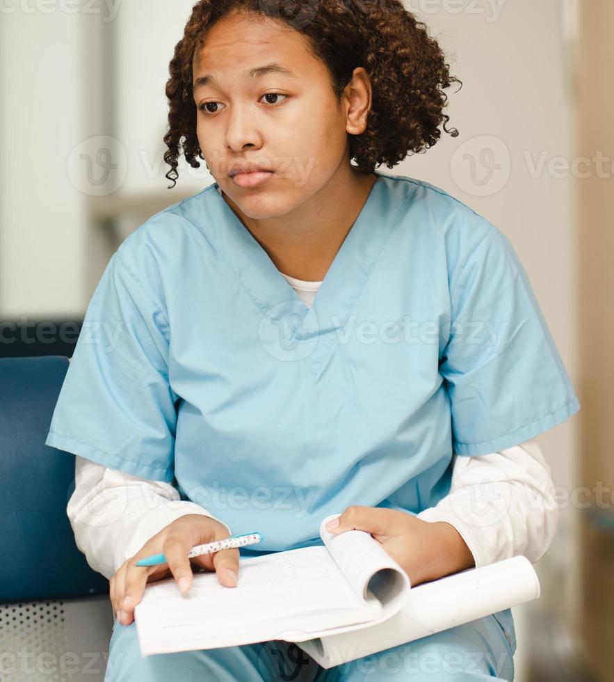 estudante de medicina cansada sentado com livro de estudo, retrato. médico interno preto de uniforme exausto de excesso de trabalho em um hospital durante a qualificação acadêmica. ocupação profissional de saúde foto