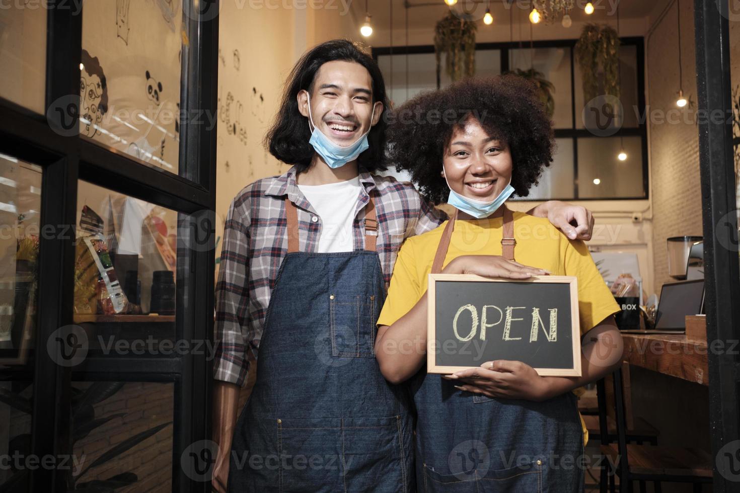 dois jovens parceiros baristas de inicialização ficam na porta do café casual, escrevem e mostram sinal aberto antes de tirar as máscaras faciais, sorrisos felizes e alegres com trabalhos de serviço de café após o estilo de vida covid-19. foto