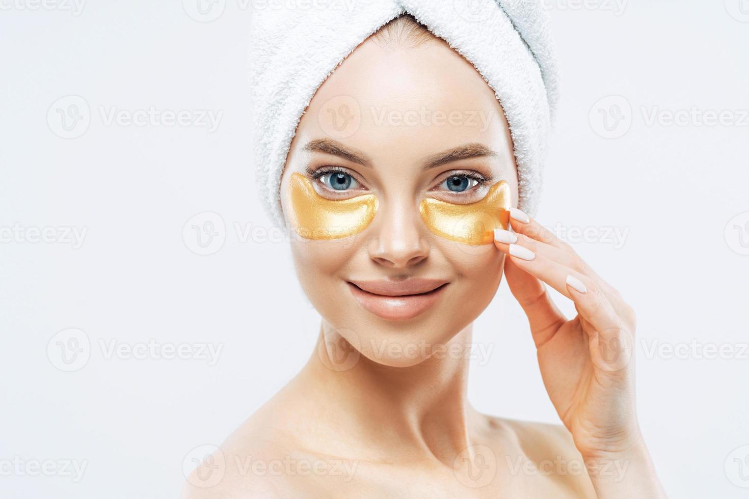 close-up tiro de mulher concurso feliz aplica manchas douradas sob os olhos, remove rugas e olheiras, tem manicure, usa toalha na cabeça, isolada sobre fundo branco. conceito de tratamento de beleza foto