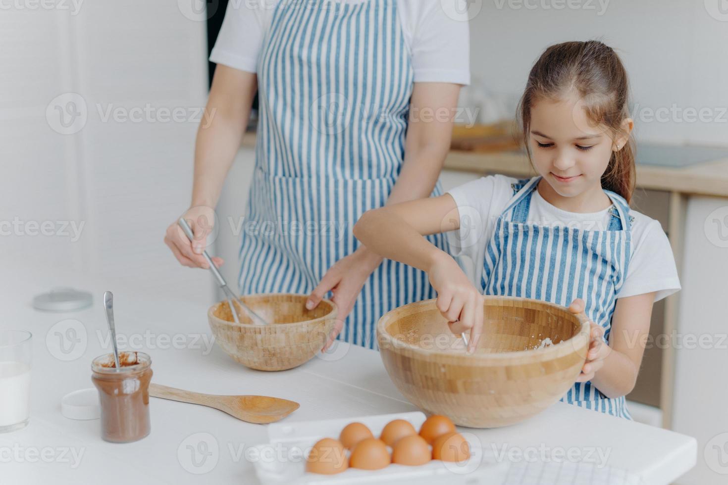 mãe dá aula de culinária para criança pequena, ficar ao lado um do outro, misturar ingredientes em grandes tigelas de madeira, fazer massa juntos, usar ovos, farinha e outros produtos. família, cozinhar, maternidade foto