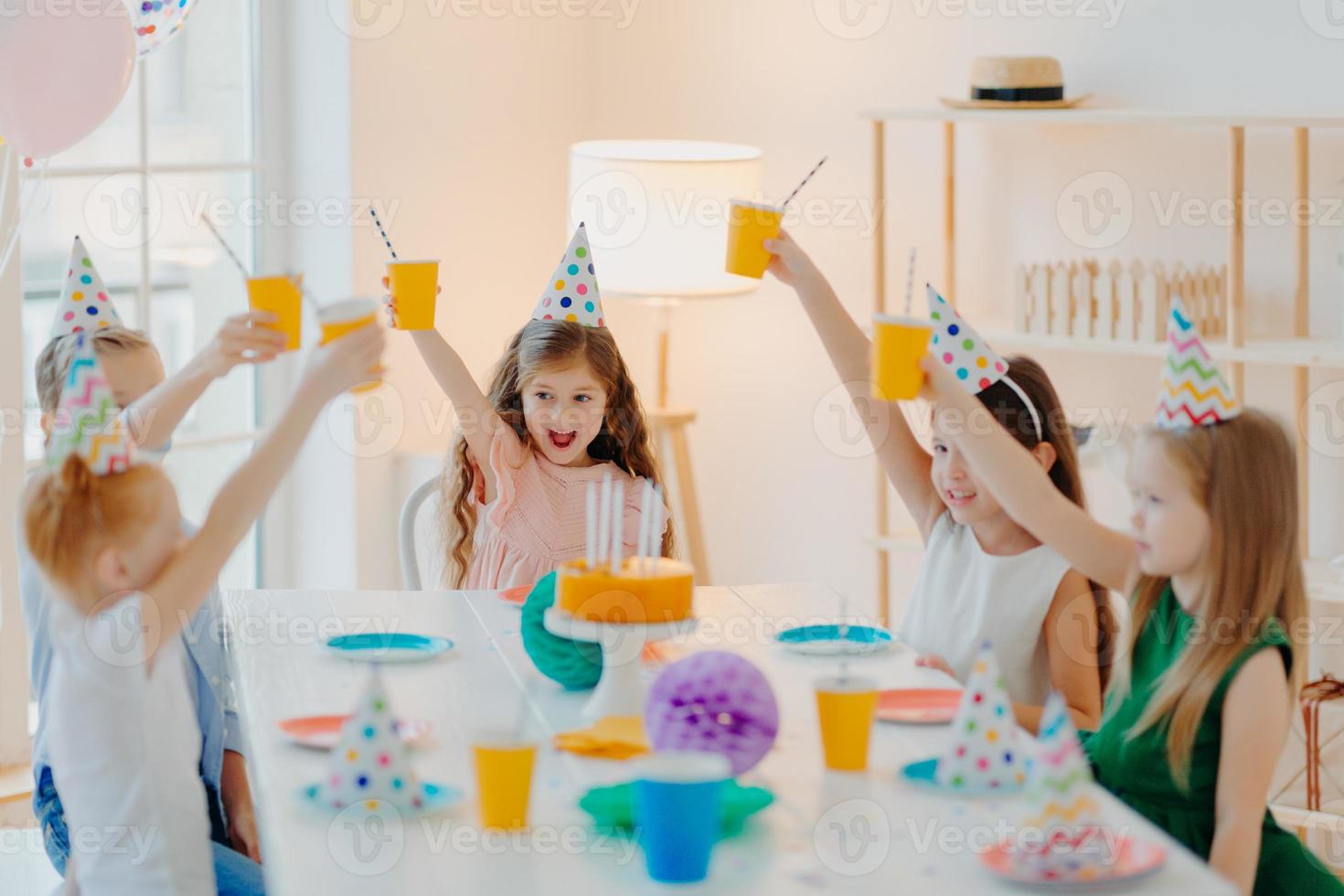grupo de crianças pré-escolares alegres comemoram aniversário juntos, se divertem, torcem com copos de bebida, usam chapéus festivos, comem bolo delicioso, sentam-se à mesa em uma sala espaçosa. crianças e conceito de festa foto