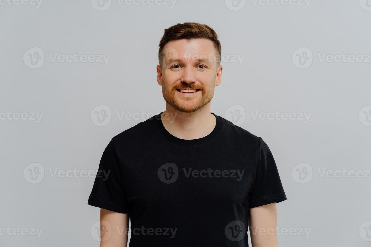 retrato de homem europeu bonito com barba por fazer vestido de camiseta preta casual sorri alegremente poses contra fundo azul expressa emoções positivas. modelo masculino feliz despreocupado de bom humor foto