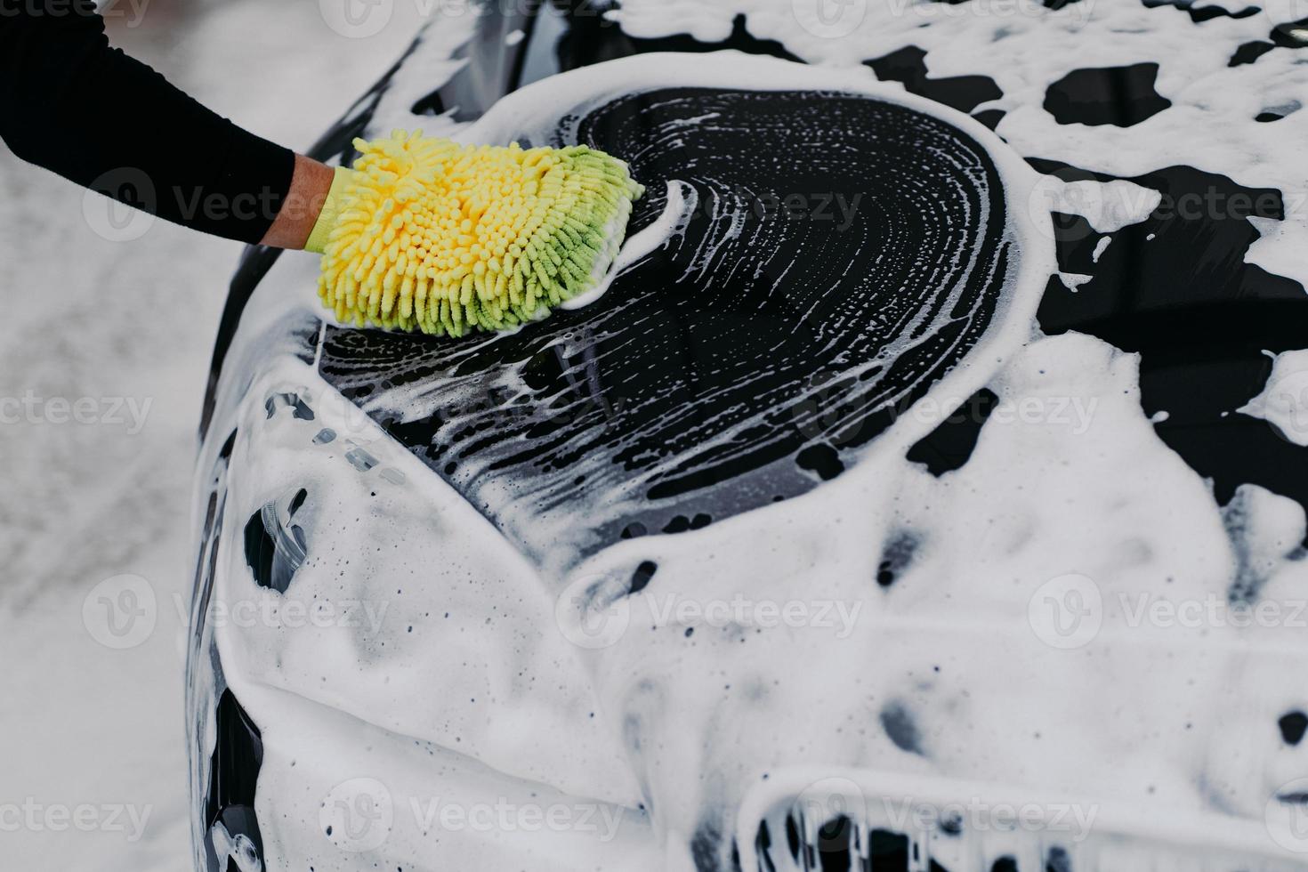 mão do homem com carro de limpeza de esfregão com xampu automático. espuma de sabão no capô preto. conceito de veículo. automóvel moderno coberto por espuma foto