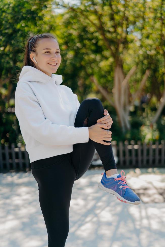 tiro vertical de mulher enérgica leva estilo de vida ativo mantém o joelho levantado exercícios ao ar livre vestido de desgaste ativo levanta perna sorrisos agradavelmente ouve faixa de áudio em fones de ouvido sem fio. treino foto