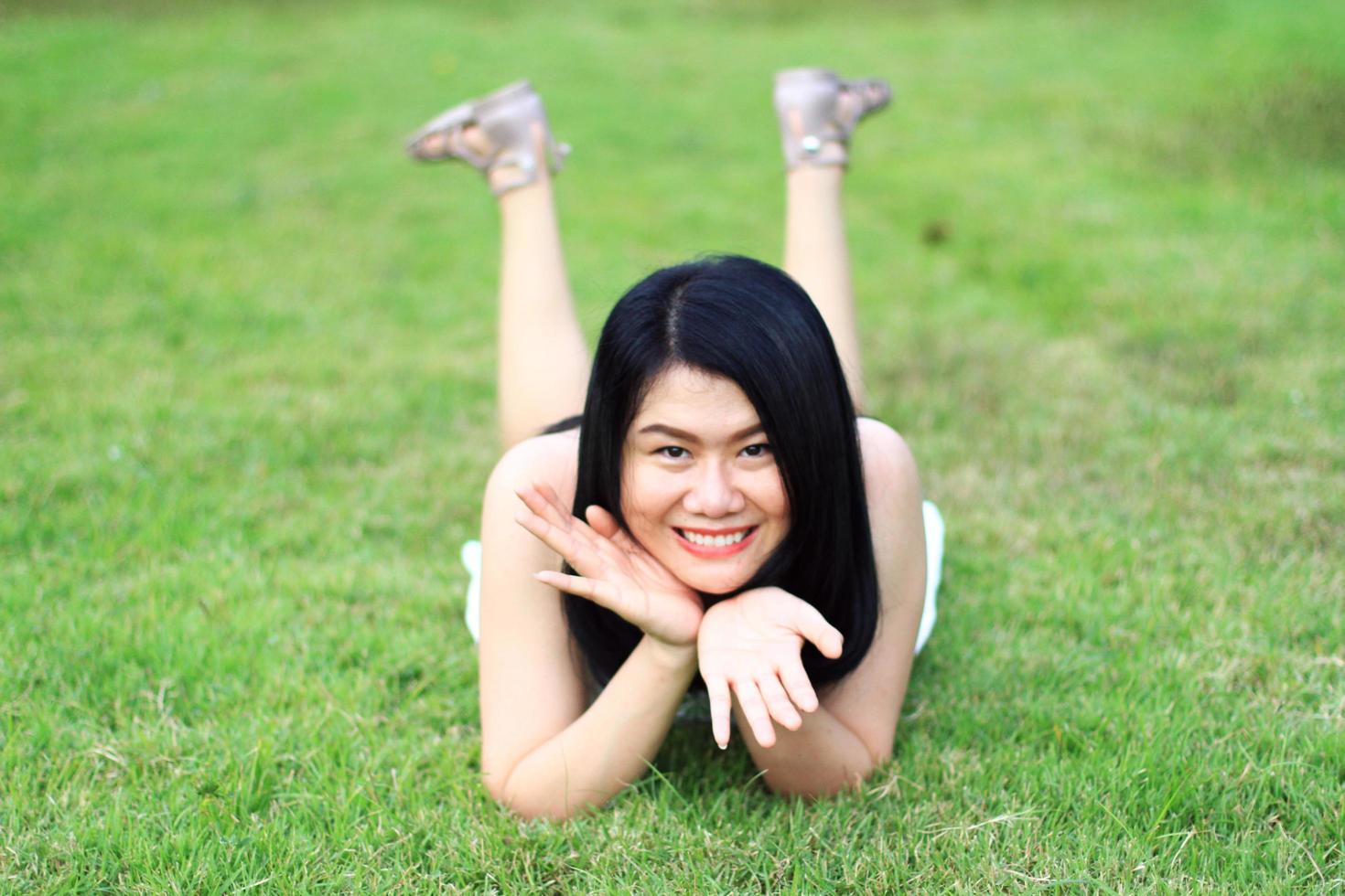 linda mulher asiática de vestido branco relaxa e sorrindo em campo verde no parque natural. jovem tailandesa desfrutar de férias com luz solar no jardim foto