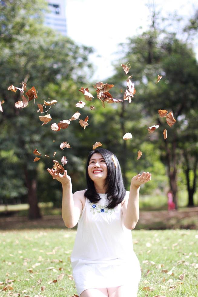 linda mulher asiática de vestido branco sorrindo e polvilhe folhas secas no parque natural. jovem tailandesa desfrutar de férias com luz solar no jardim foto