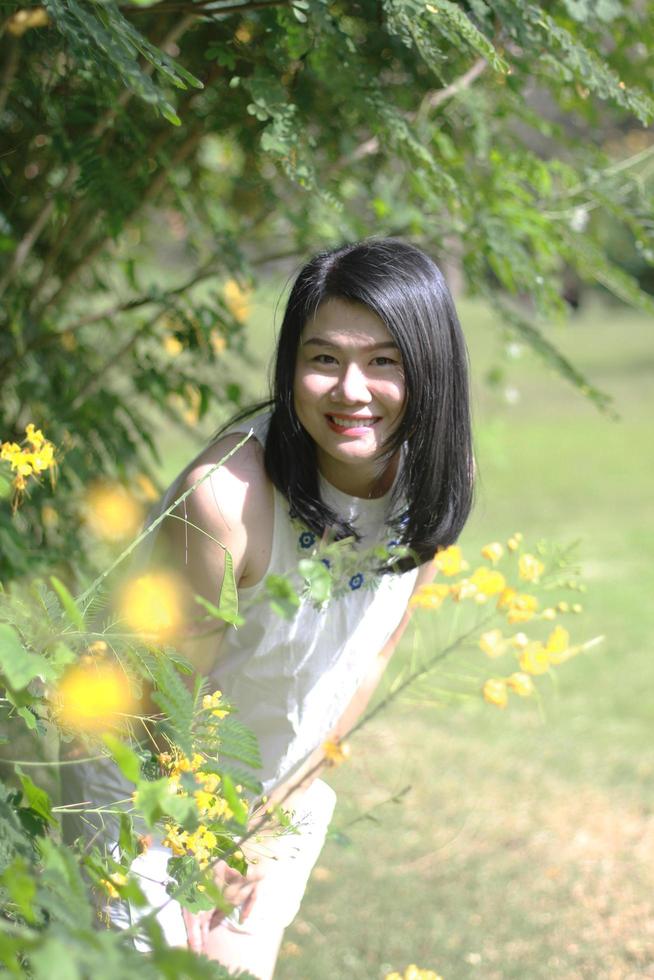 linda mulher asiática de vestido branco relaxa e cheirando flores no jardim natural. jovem tailandesa desfrutar de férias com luz solar no parque floral amarelo foto