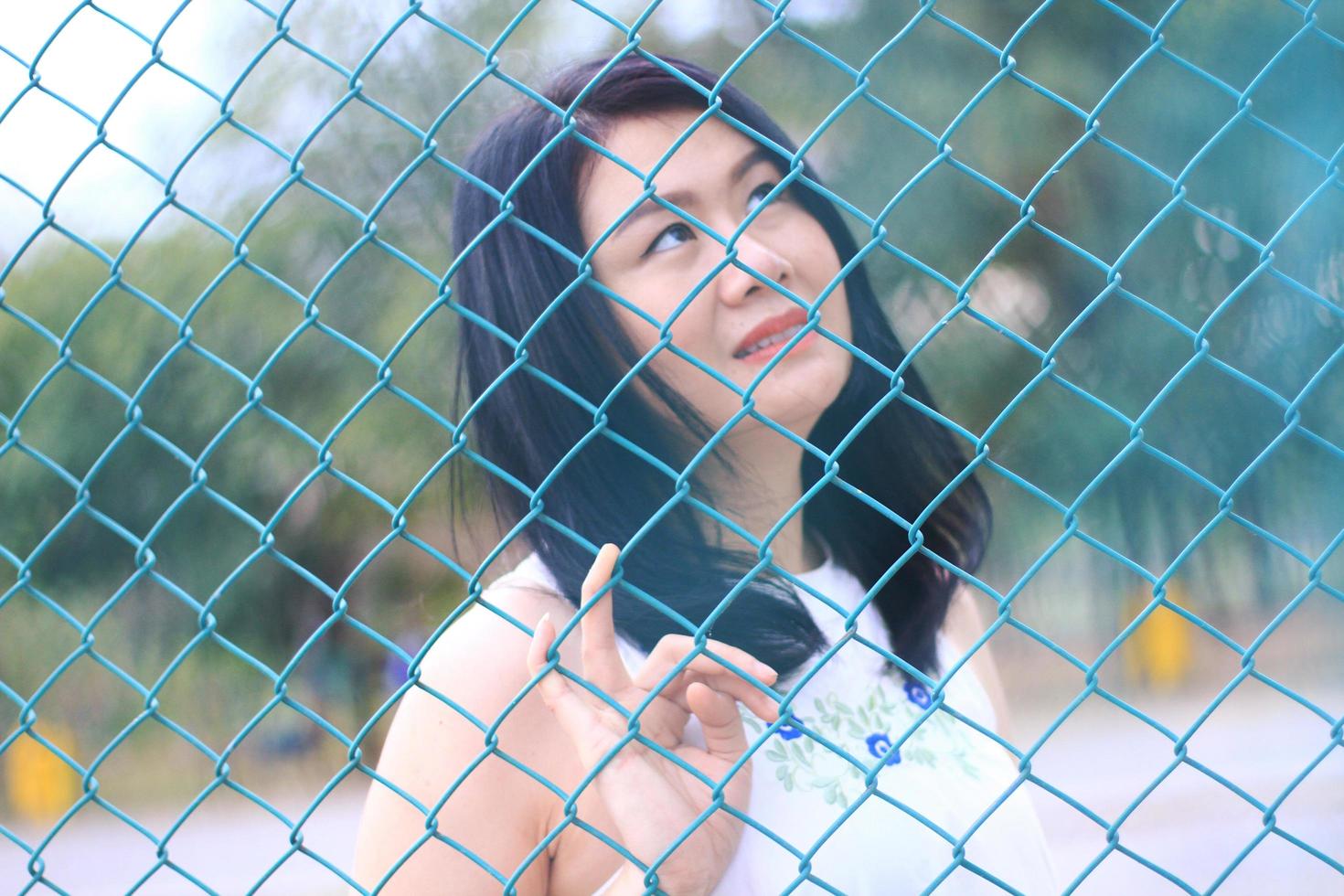 linda mulher asiática de vestido branco sentado e sorrindo no parque natural. menina tailandesa ou menina chinesa desfrutar de férias com luz solar no jardim. fechou o foco na cerca de aço ralar azul. foto