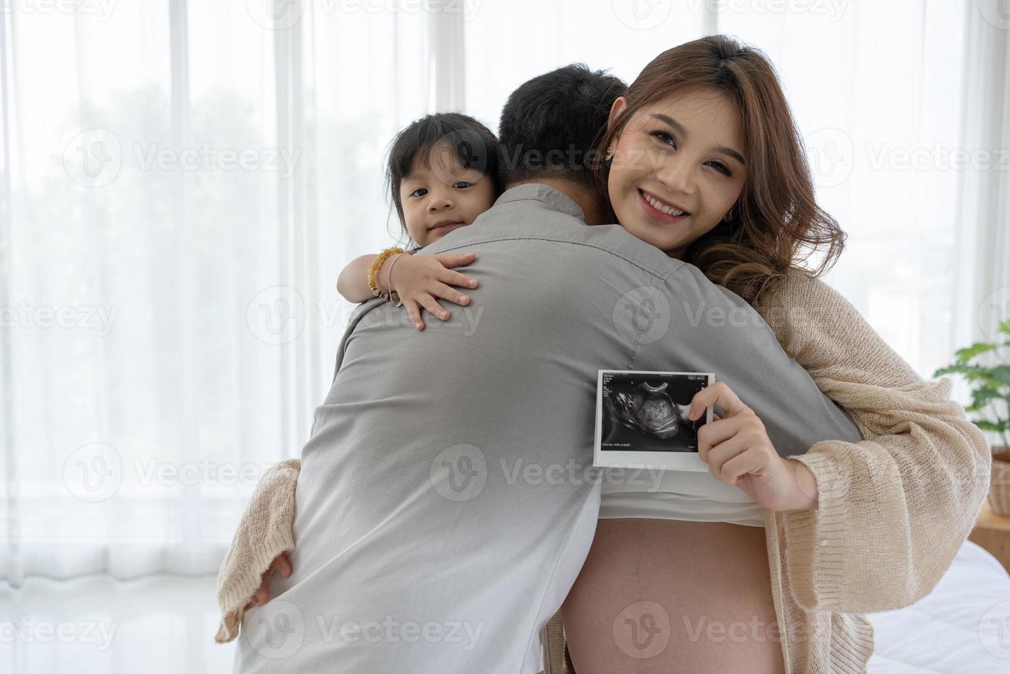 família grávida abraçando e em pé perto da janela. esposa segurando ultra-sonografia do bebê foto