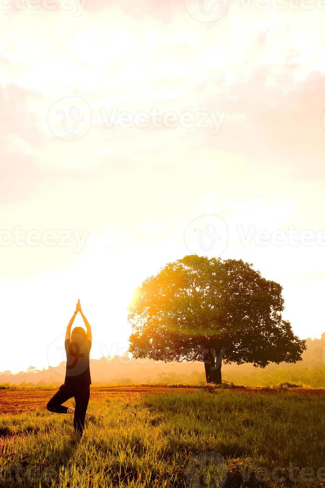 a mulher pratica ioga no prado com árvore e nascer do sol ao fundo foto