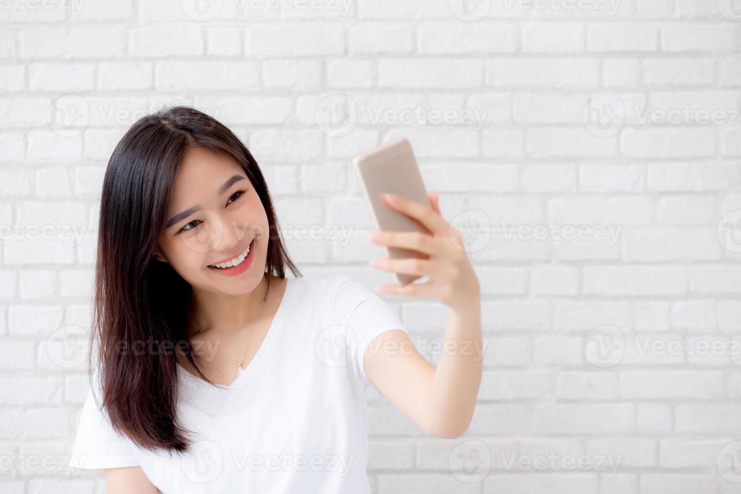 belo retrato jovem mulher asiática tomando uma selfie com telefone móvel inteligente no fundo branco de cimento concreto, a garota está fotografando com feliz e sorri com diversão, conceito de estilo de vida. foto