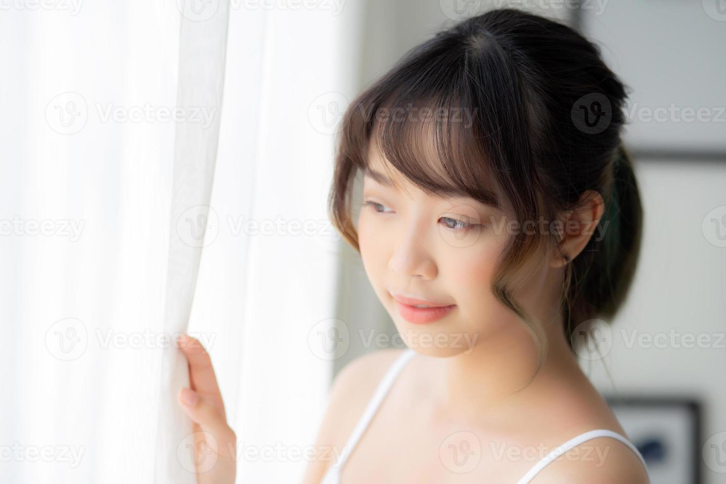 retrato de uma bela jovem asiática em pé olhe para a janela e sorria enquanto acorda com a luz do sol de manhã, menina feliz com estilo de vida fresco e alegre, e relaxe para o conceito de bem-estar. foto