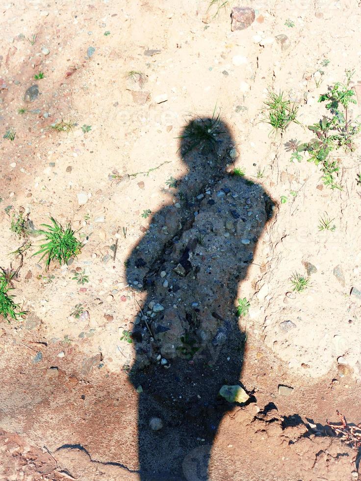 longa sombra de uma pessoa de casaco com capuz na areia seca com grama verde crescente e muitas pedras e rochas foto