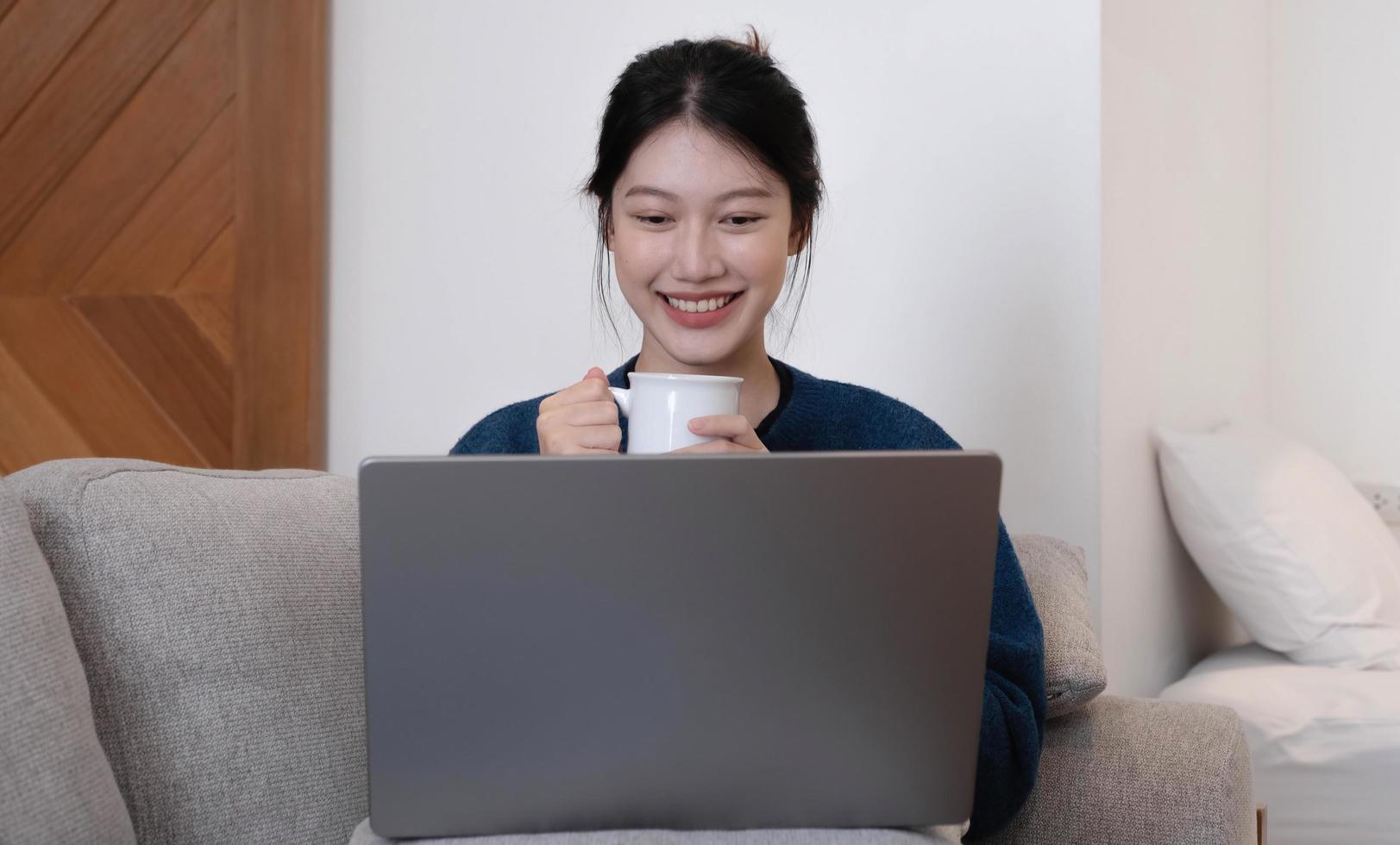 menina asiática trabalhando no laptop online, usando a internet e segure a taça. sentado no sofá em casa. foto