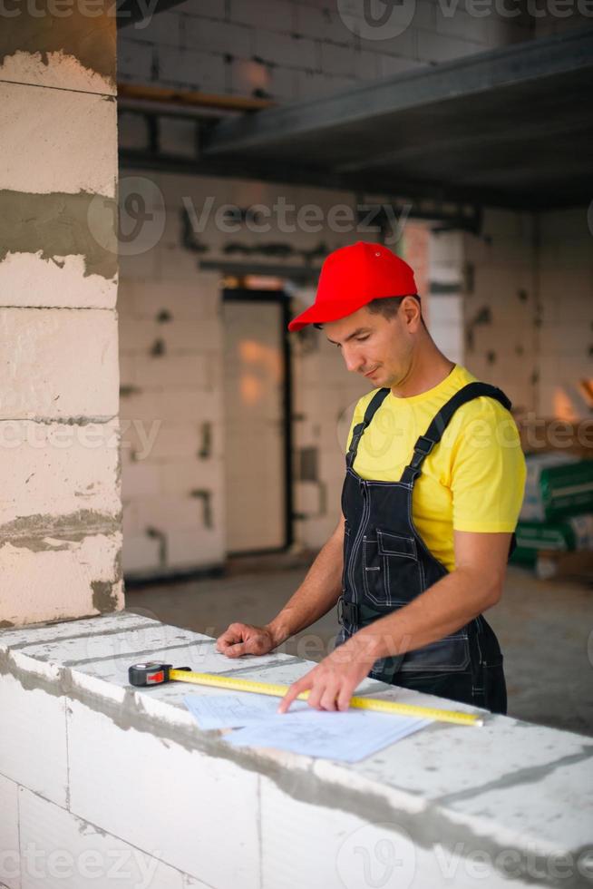 trabalhador da construção civil no canteiro de obras mede o comprimento da abertura da janela e a parede de tijolos com fita métrica. cottage são feitos de blocos de concreto porosos, roupas de trabalho - macacão e boné de beisebol foto