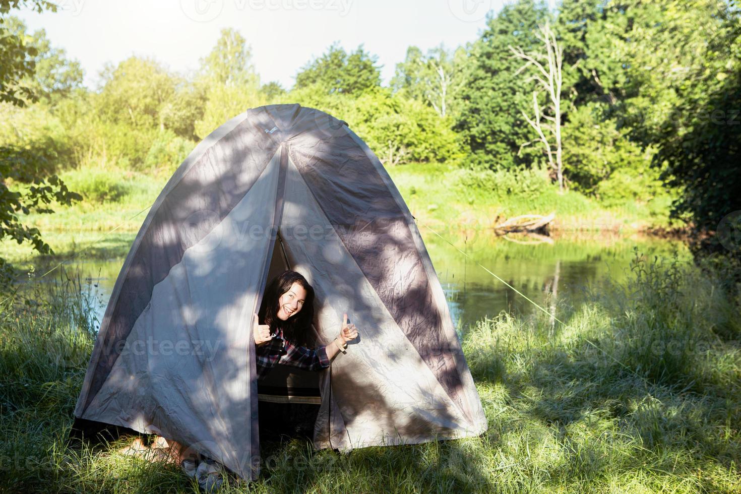 uma mulher feliz em uma camisa xadrez parece fora de uma tenda turística em uma caminhada na margem do rio pela manhã. acampar na natureza, pernoite na natureza, férias em família e aventuras. foto