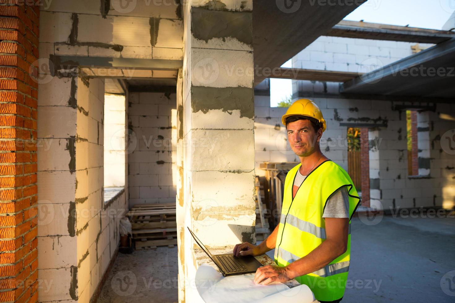 engenheiro de construção no canteiro de obras de uma casa feita de blocos de concreto porosos trabalha em um computador em um colete de segurança reflexivo e capacete. projeto, construção, projeto de desenhos, cheque foto