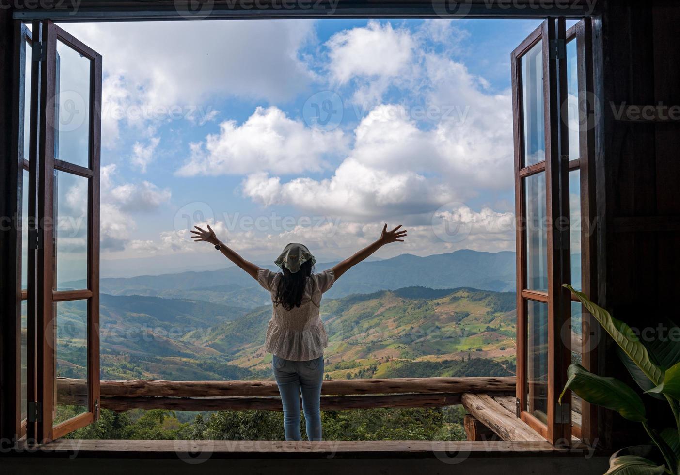 parte traseira da mulher asiática com os braços da liberdade abertos em pé atrás da grande janela de madeira com vista para a montanha e céu azul nublado em doi chang, atração de viagens em chiang rai foto