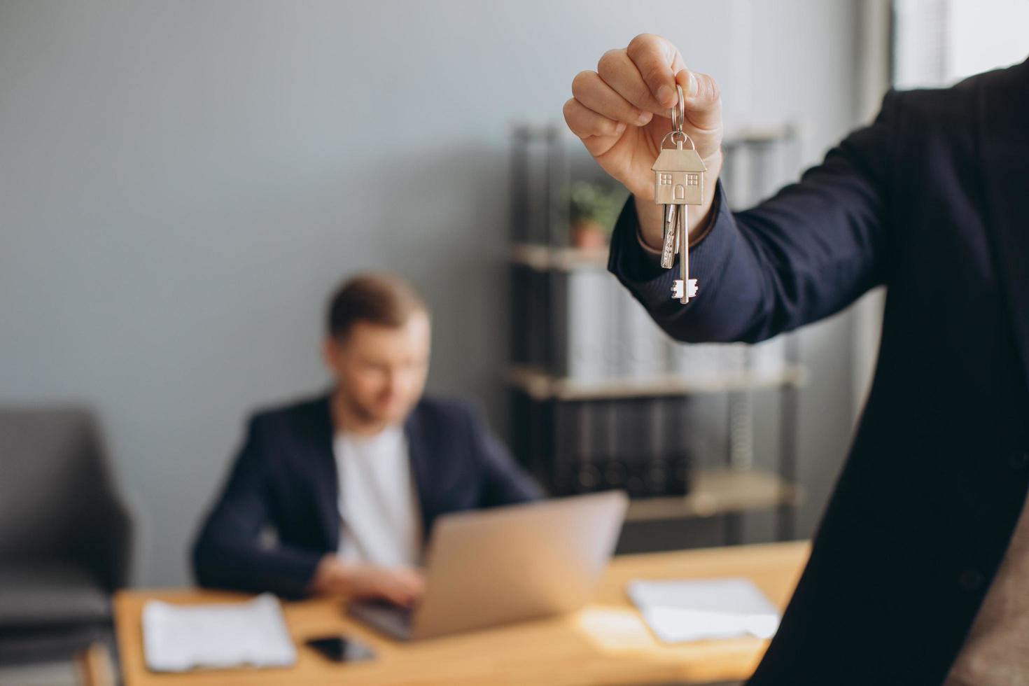 sorrindo jovem moderno de terno detém as chaves do apartamento comprado no fundo do corretor de imóveis e escritório foto