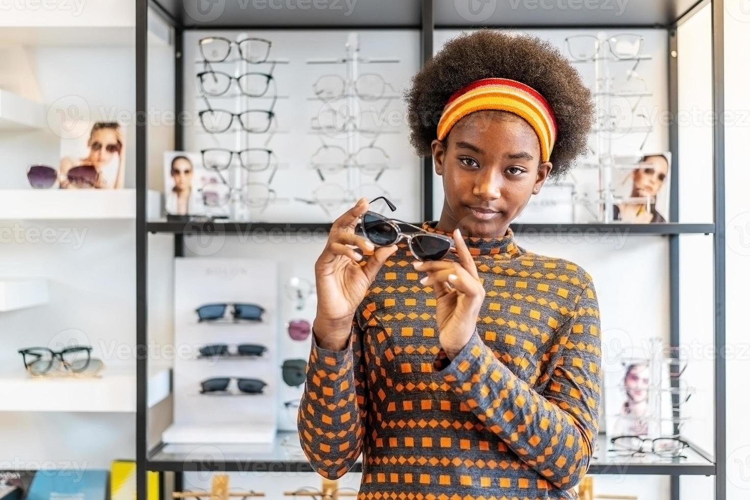 precisa de um novo óculos afro-americano de mulher jovem posando com óculos novos em pé na loja de óptica. conceito de oftalmologista moderno foto