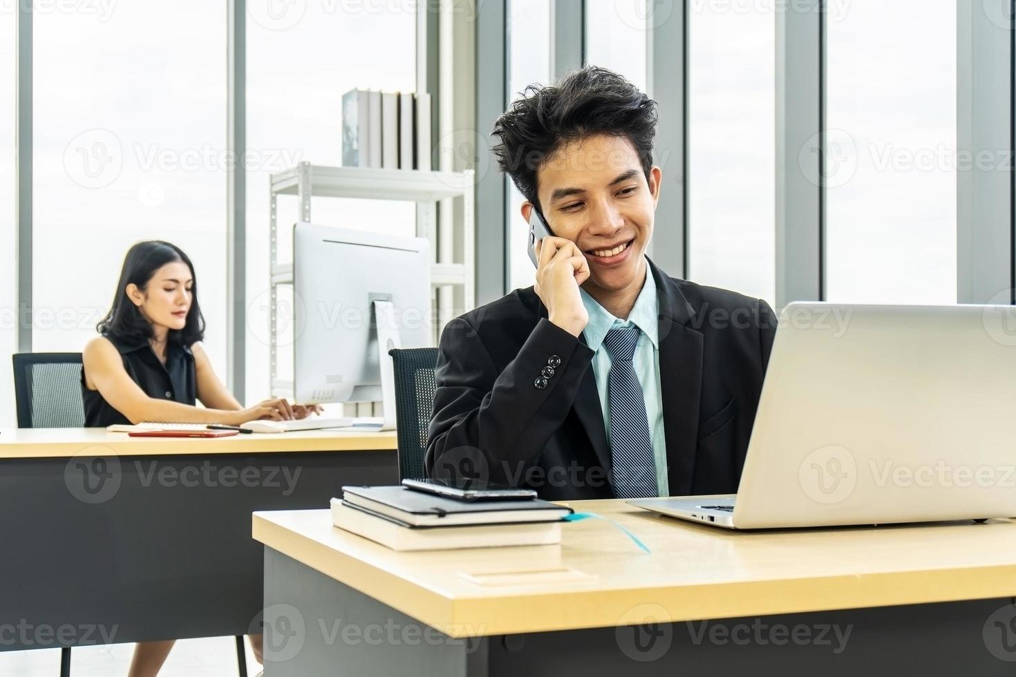 jovem empresário sentado no escritório falando no celular, sentado na mesa, usando laptop, sorrindo, empresária trabalhando no laptop em sua estação de trabalho em segundo plano. foto