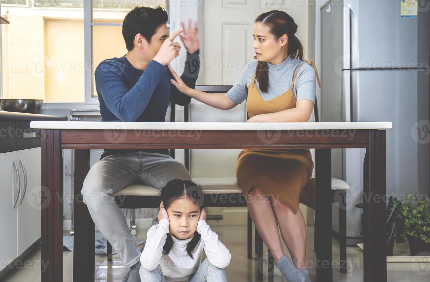 filhinha asiática triste e entupir os ouvidos debaixo da mesa enquanto os pais brigam na cozinha, design de fotos para problemas familiares e conceito infeliz