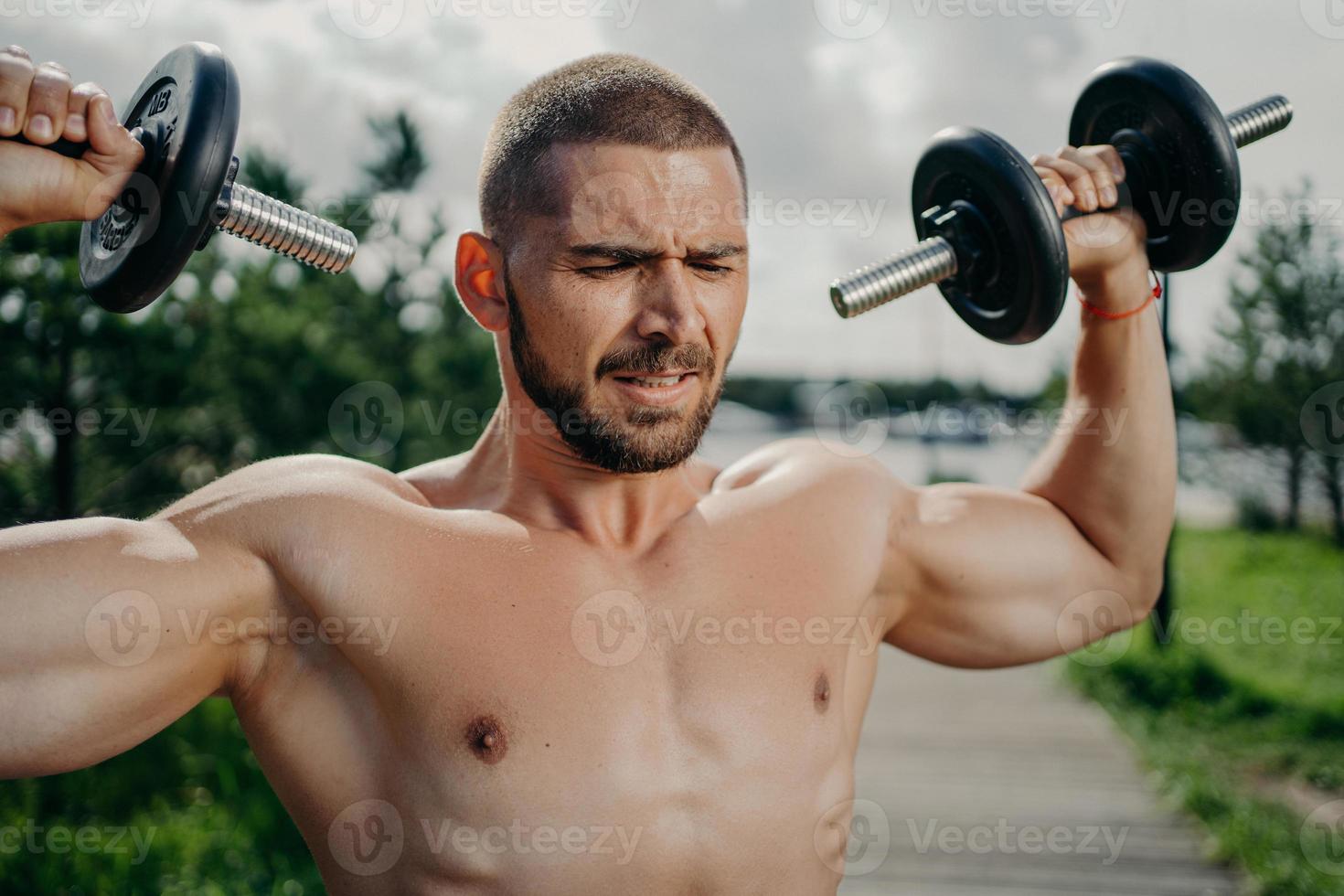 desportivo determinado musculoso homem europeu exercsisses com halteres, faz levantamento de peso ao ar livre, tem torso nu, treina músculos, quer ter corpo atlético. conceito de força e motivação. foto