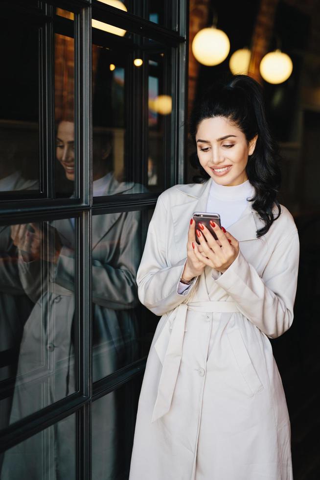 elegante bela jovem morena com rabo de cavalo vestido com capa de chuva branca com rosto agradável e manicure vermelha perfeita segurando smartphone olhando para a tela com um sorriso agradável foto