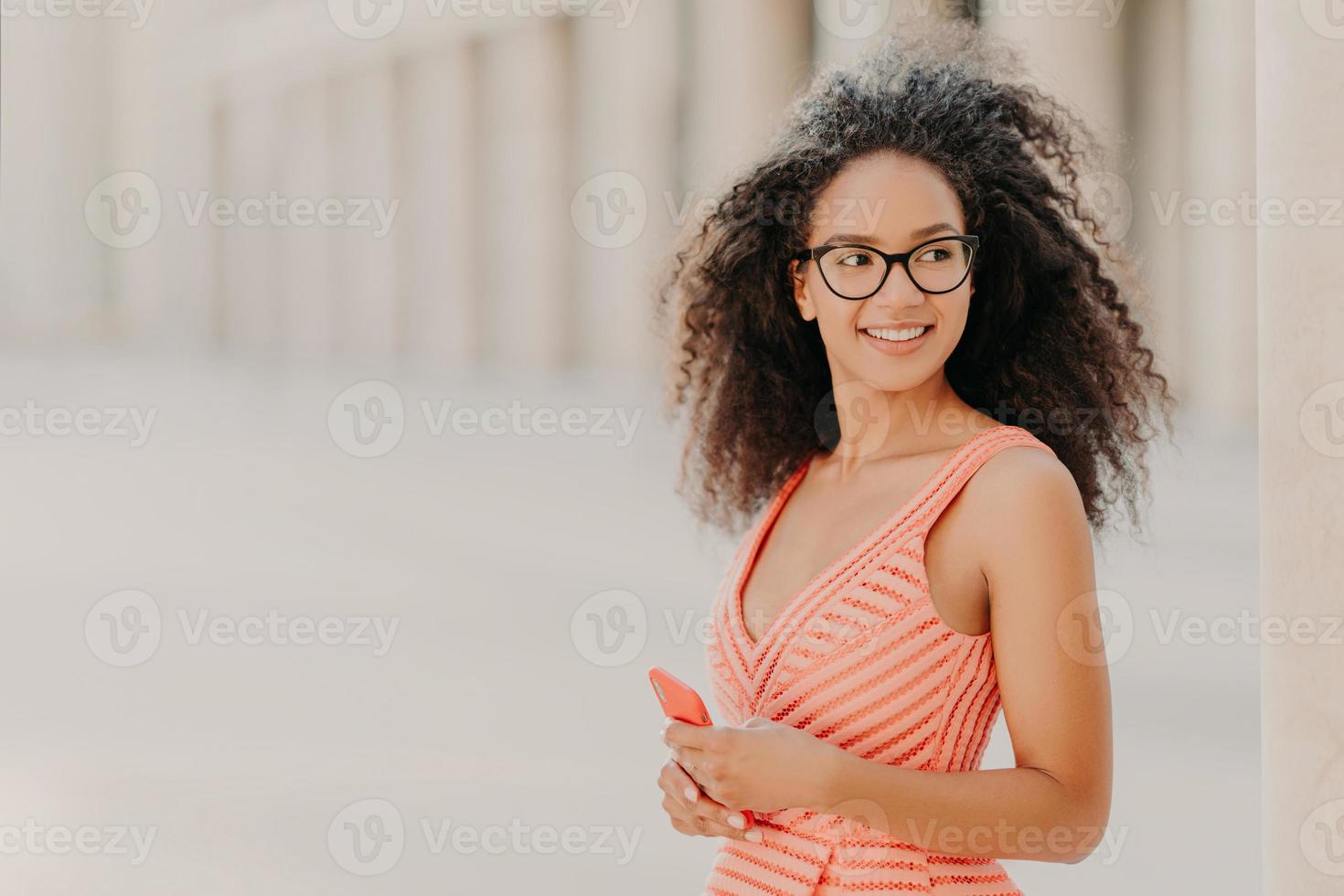 menina milenar satisfeita com cabelo encaracolado, usa vestido rosa, óculos ópticos, focado de lado com sorriso, fica ao ar livre, espera por ligação, gosta de conexão de internet de alta velocidade, atualiza novo aplicativo foto