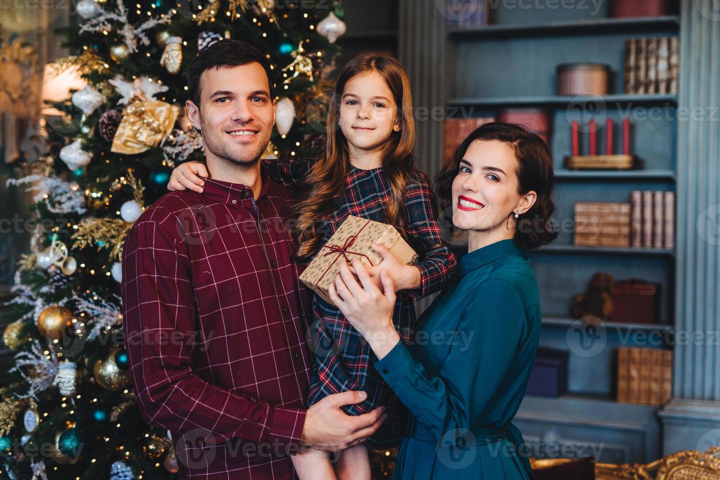 família feliz fica junto contra abeto decorado, olha alegremente para a câmera, segura o presente, comemora o ano novo no círculo familiar. menina feliz em passar férias com a mãe e o pai foto