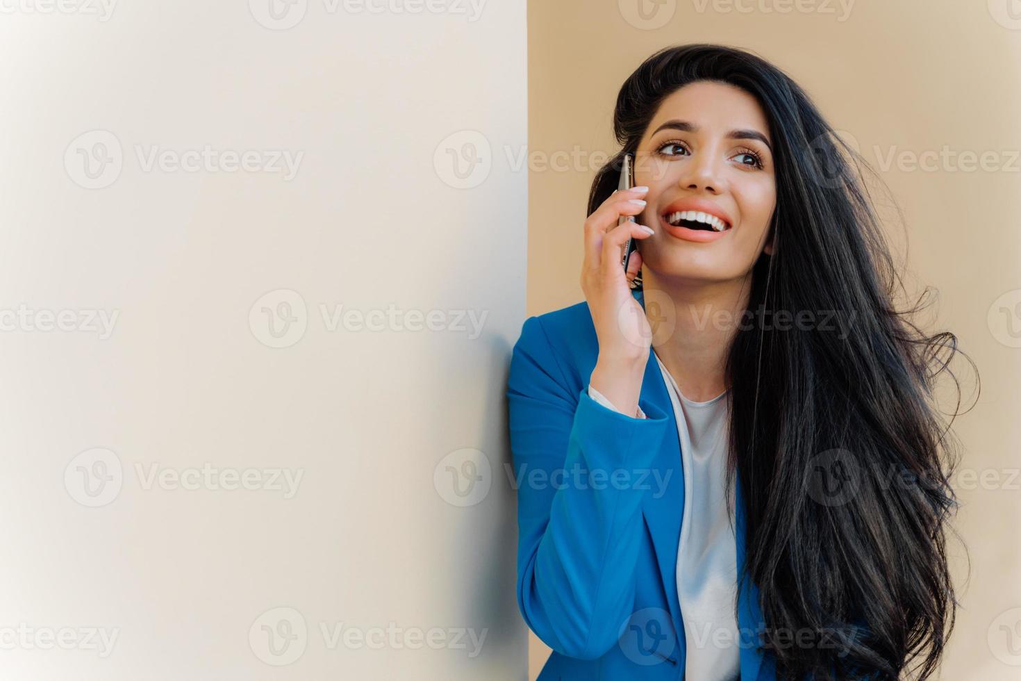 jovem empresária positiva sorri alegremente, se comunica no celular, vestida formalmente, focada em algum lugar, tem maquiagem mínima. alegre empreendedora resolve problemas de trabalho de bom humor foto