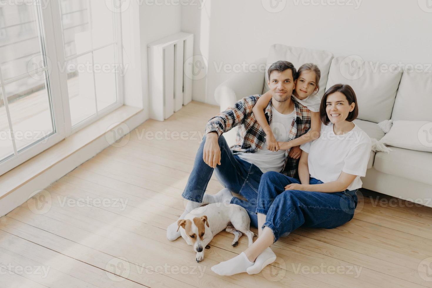 adorável família europeia de pai, mãe e filha sentam-se no chão perto do sofá na espaçosa sala branca, cachorro com pedigree fica perto, tem dia de mudança, muda de local de vida, expressa felicidade. foto