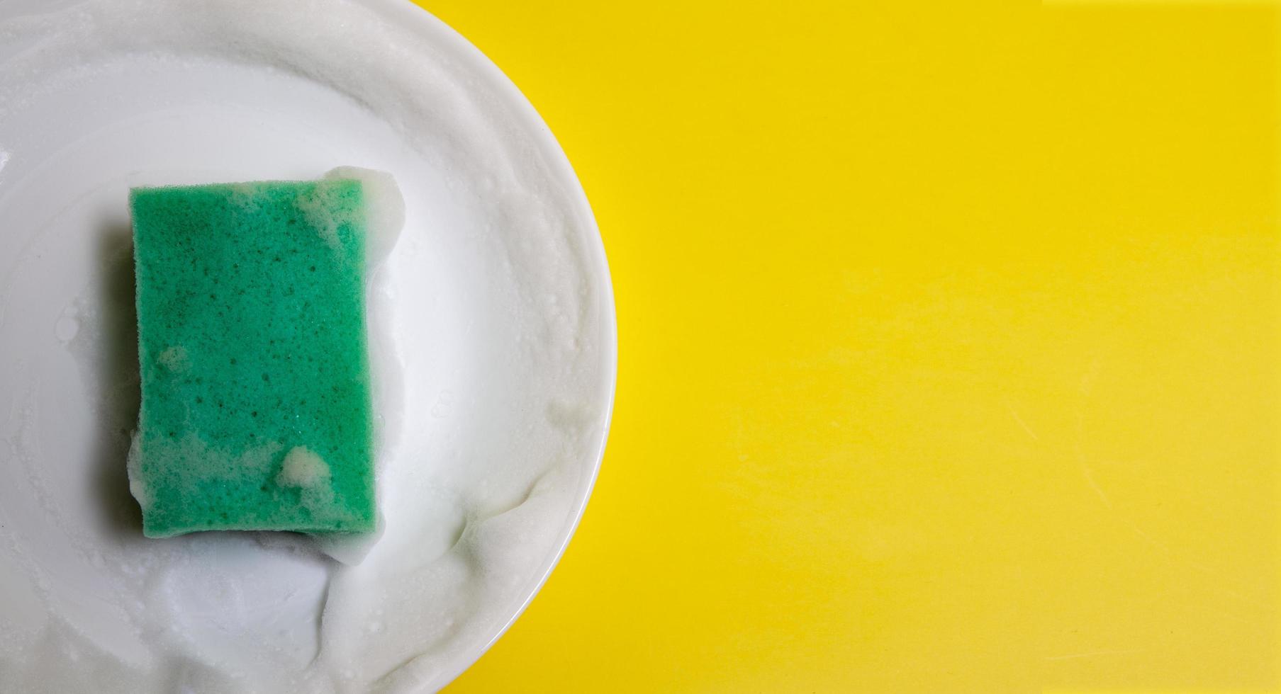 esponja verde em espuma em um prato branco sobre um fundo amarelo. conceito de lavar pratos foto
