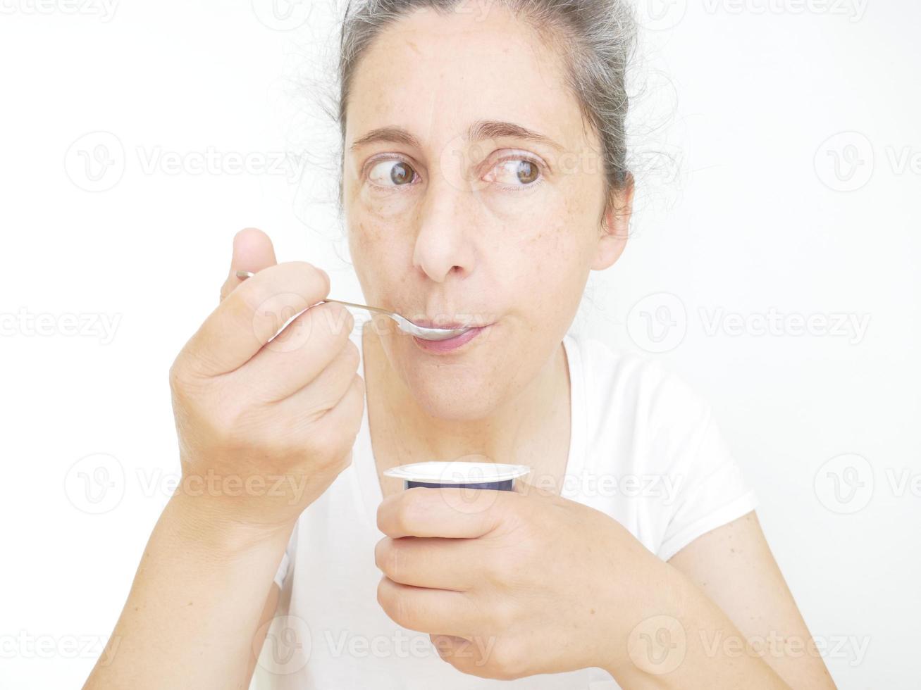 mulher de quarenta e nove anos em uma camiseta branca contra um fundo branco comendo um iogurte foto