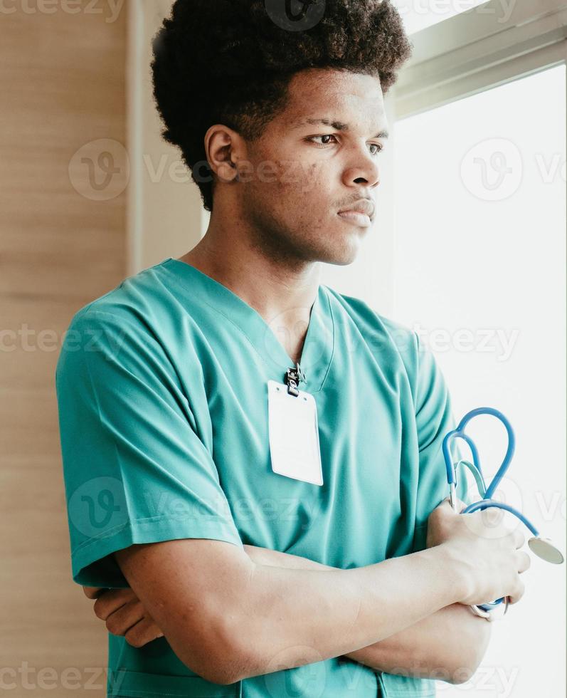 pensativo jovem médico americano africano em pé pela janela olhando para longe os braços cruzados. médico masculino retrato detém estetoscópio pensando seriamente, ponderando. estilo de vida de ocupação médica profissional. foto