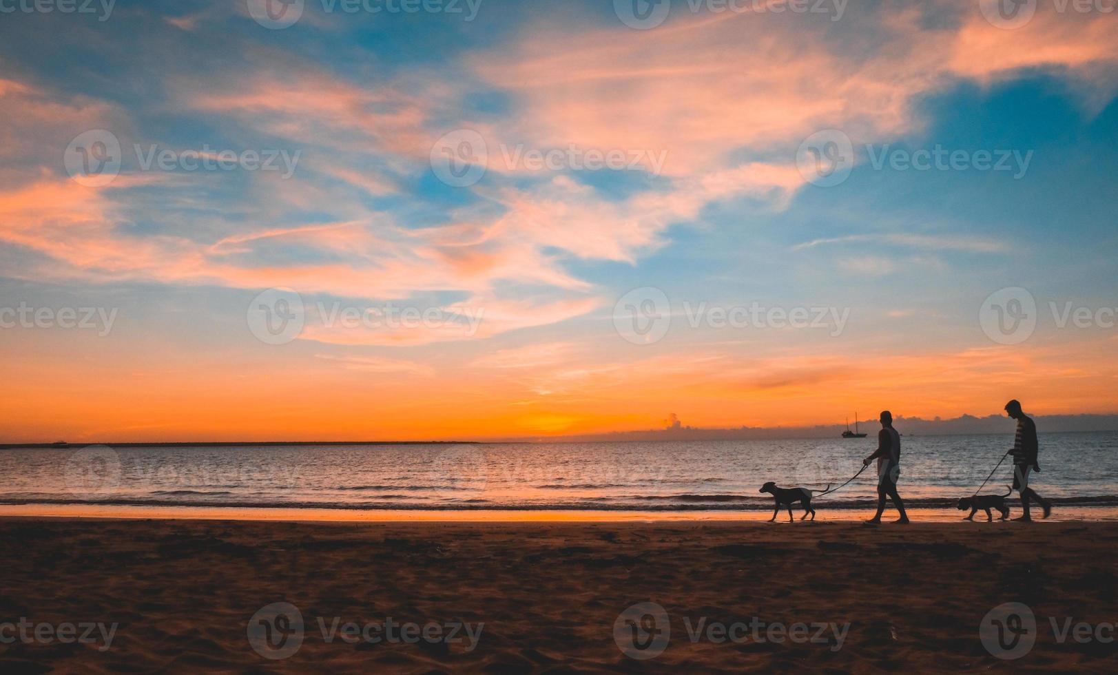 silhueta de pessoas passeando com seus cães na praia durante o pôr do sol. céu azul e nuvens pitorescas. foto