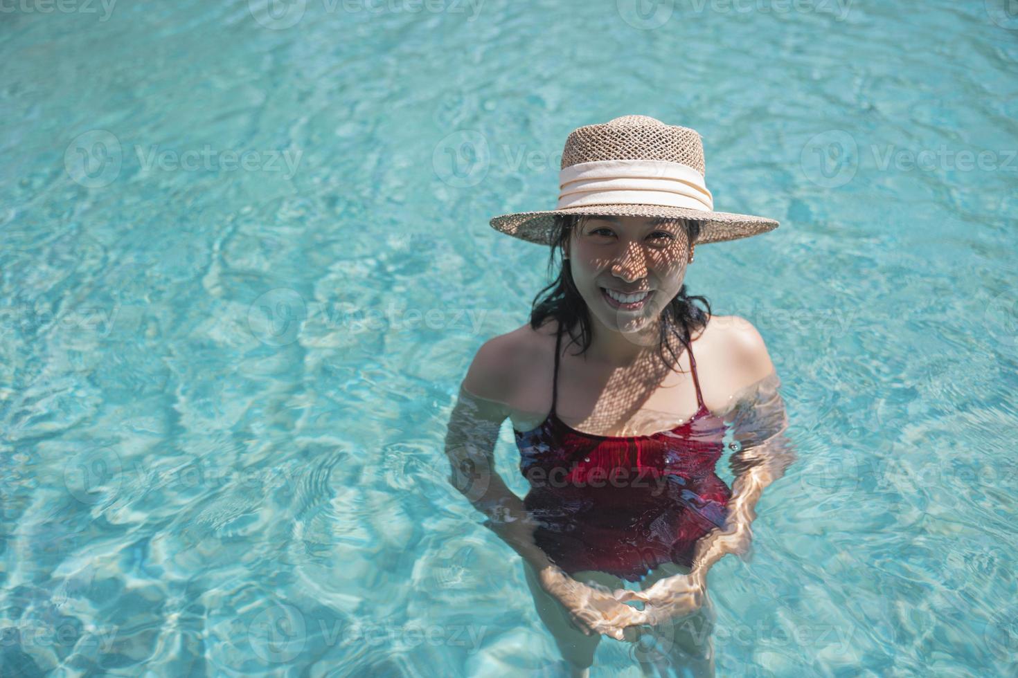 feliz mulher asiática em maiô vermelho e um chapéu de palha relaxante na piscina olhando para a câmera à beira da piscina em koh mak, phangnga, tailândia. relaxe e viaje o conceito. férias de verão resort conforto foto